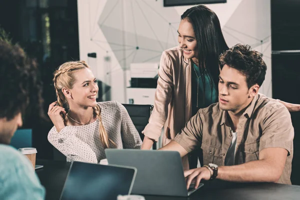 Colegas de negocios multiculturales que trabajan y hablan en la mesa con ordenadores portátiles en la oficina moderna - foto de stock