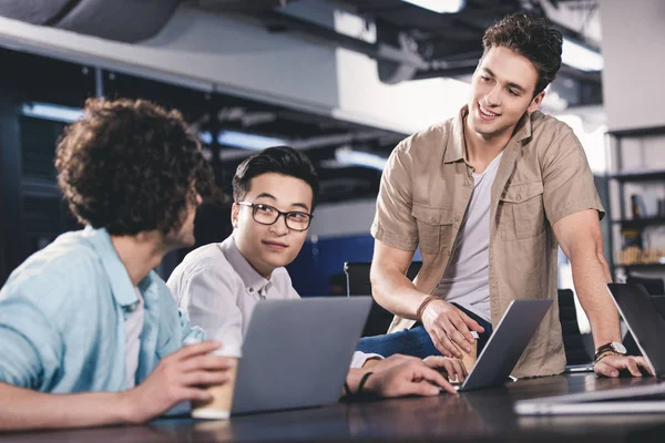 Three multicultural businessmen having meeting with paper cups of coffee at table with laptops in modern office — Stock Photo