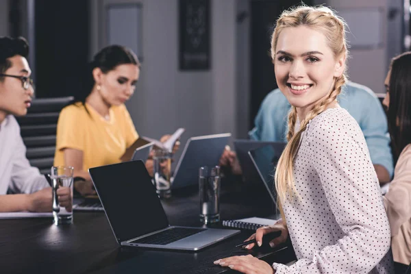 Jeune femme d'affaires souriante regardant la caméra tandis que ses collègues travaillant derrière au bureau moderne — Photo de stock