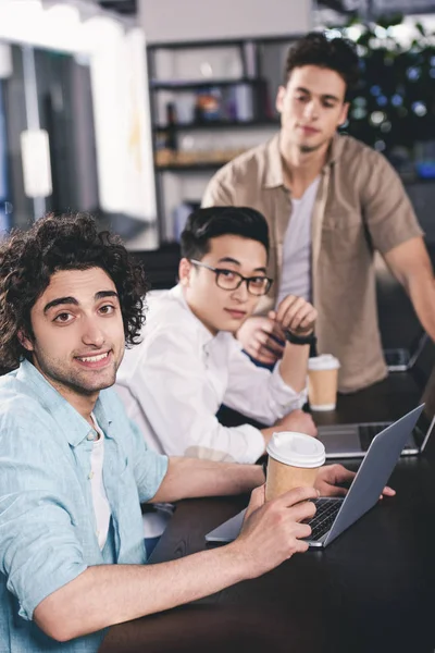 Three multiethnic businessmen with paper coffee cups sitting at table with laptops at modern office — Stock Photo