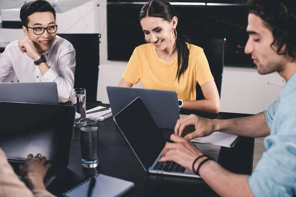 Sorrindo grupo multicultural de parceiro de negócios conversando à mesa com laptops no escritório moderno — Fotografia de Stock