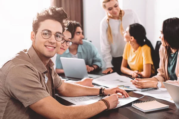 Dos hombres de negocios multiculturales en gafas mirando a la cámara mientras sus socios discuten detrás en la oficina moderna - foto de stock