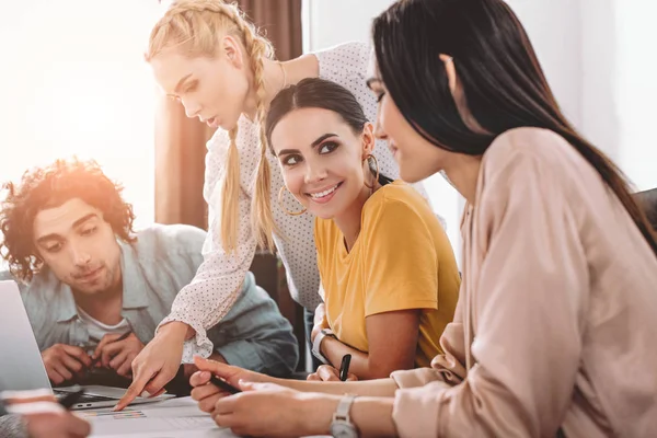 Young businesswoman pointing by finger on graphs to male colleague while two businesswomen talking to each other at modern office — Stock Photo