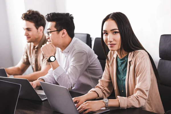 Jovem asiático empresária digitação no laptop enquanto ela colegas tendo discussão no moderno escritório — Fotografia de Stock
