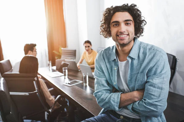 Lächelnder junger Geschäftsmann mit verschränkten Händen, der in die Kamera schaut und seine Partner, die im modernen Büro hinter ihm arbeiten — Stockfoto