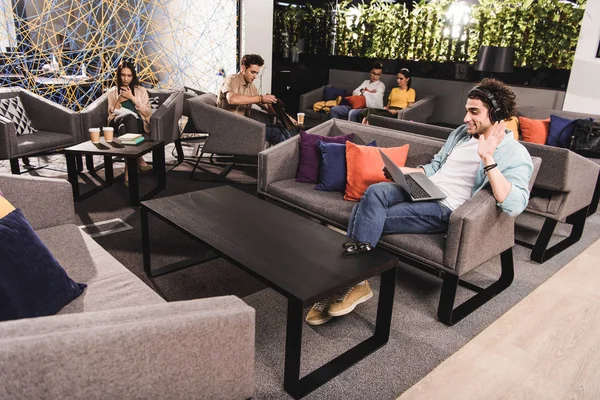 Junger Geschäftsmann mit Kopfhörer sitzt auf Couch mit Laptop und winkt von Hand in modernem Coworking-Büro — Stockfoto
