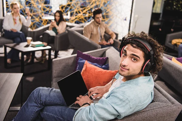 Joven hombre de negocios en auriculares sentados en el sofá y el uso de la computadora portátil con pantalla en blanco en la moderna oficina de coworking - foto de stock