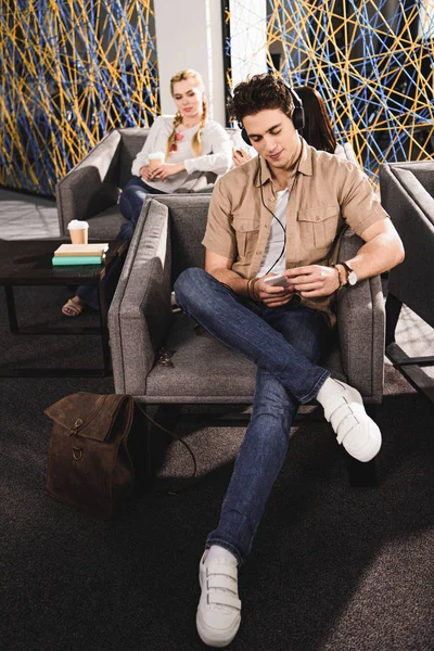 Young businessman in headphones with smartphone sitting on armchair and businesswoman sitting behind with coffee at modern coworking office — Stock Photo