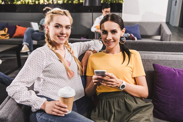 Two smiling businesswomen with coffee and smartphone at modern coworking office — Stock Photo