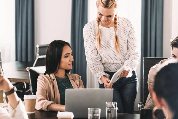 Sonriente mujer de negocios mostrando digital tablet a asiático mujer colega mientras otros socios tener discusión en moderno oficina - foto de stock