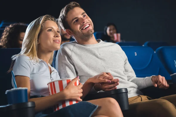 Smiling couple with popcorn and soda drink holding hands while watching film together in cinema — Stock Photo