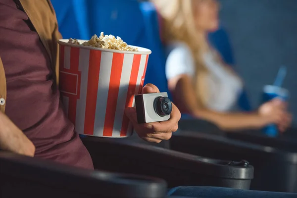 Vista parcial del hombre con una pequeña cámara de video y palomitas de maíz filmando ilegalmente una película en el cine - foto de stock