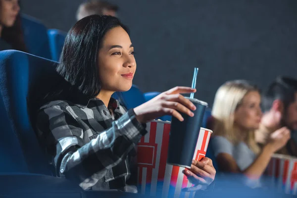 Vista lateral de sonriente mujer asiática con palomitas de maíz y soda bebida viendo película en cine - foto de stock