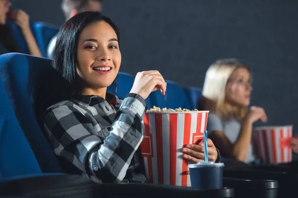 Улыбающаяся азиатка с попкорном, смотрящая на камеру в кино — стоковое фото
