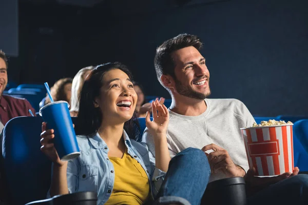 Feliz pareja interracial con palomitas de maíz y refrescos viendo películas juntos en el cine - foto de stock