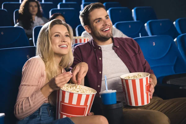 Улыбающаяся пара с попкорном и напитком, смотрящая фильм вместе в кино — стоковое фото