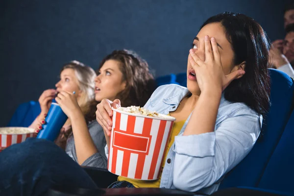 Mujeres multirraciales enfocadas y asustadas con palomitas de maíz viendo películas juntas en el cine - foto de stock