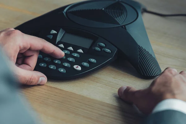 Cropped shot of businessman pushing button of conference phone on wooden table at office — Stock Photo