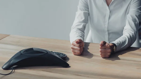 Cropped shot of businesswoman in white shirt sitting in front of conference phone on table and making fists — Stock Photo