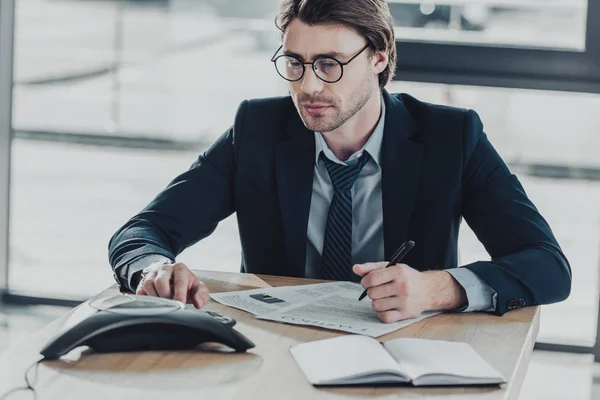Schöner junger Geschäftsmann mit viel Papierkram mit Konferenztelefon im Büro — Stockfoto