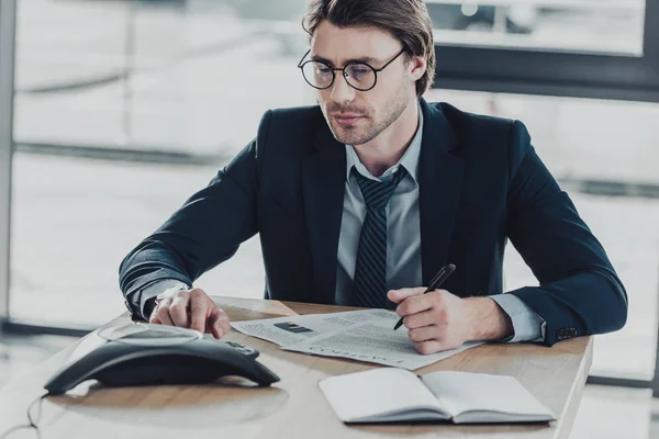 Bonito jovem empresário com muita papelada usando telefone conferência no escritório — Fotografia de Stock