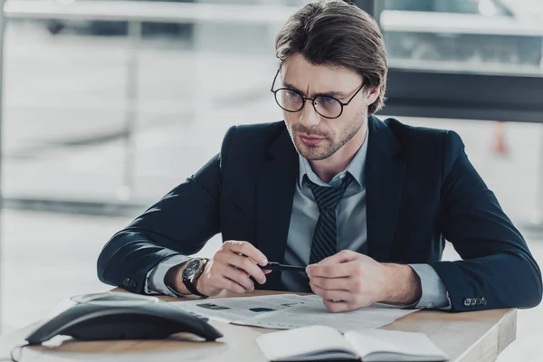 Jeune homme d'affaires sérieux avec divers documents sur la table regardant la conférence — Photo de stock