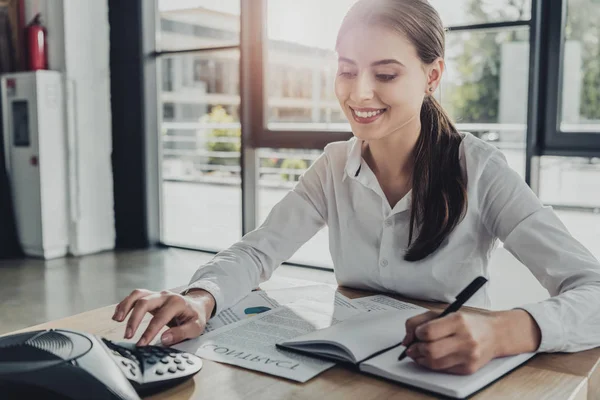 Young smiling businesswoman doing paperwork and pushing button of conference phone on table at office — Stock Photo