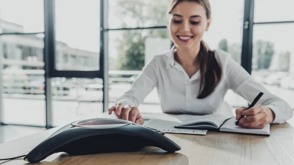 Joven feliz mujer de negocios haciendo papeleo y pulsando el botón de teléfono de la conferencia en la mesa en la oficina - foto de stock