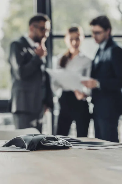 Nahaufnahme eines Konferenztelefons mit verschwommenen Geschäftsleuten, die im Hintergrund im modernen Büro zusammenarbeiten — Stockfoto