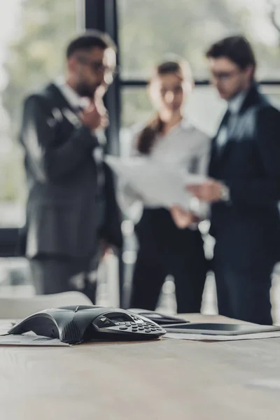 Plan rapproché du téléphone de conférence avec des gens d'affaires flous travaillant ensemble sur le fond au bureau moderne — Photo de stock