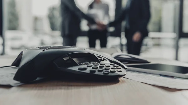 Close-up shot of conference phone with blurred business people shaking hands on background at modern office — Stock Photo