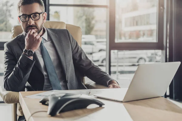 Apuesto empresario reflexivo sentado en el lugar de trabajo y mirando hacia otro lado - foto de stock