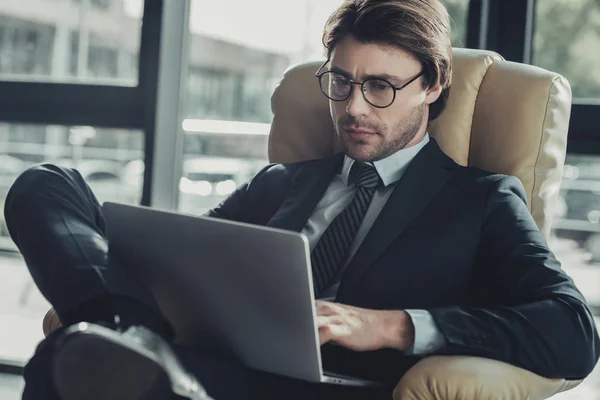 Красивий концентрований бізнесмен сидить у кріслі і використовує ноутбук — стокове фото