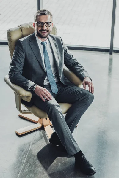 Vue grand angle de bel homme d'affaires heureux assis sur un fauteuil de luxe et regardant la caméra au bureau moderne — Photo de stock