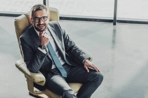 Visão de alto ângulo de belo homem de negócios sorridente sentado em poltrona de luxo no escritório moderno — Fotografia de Stock