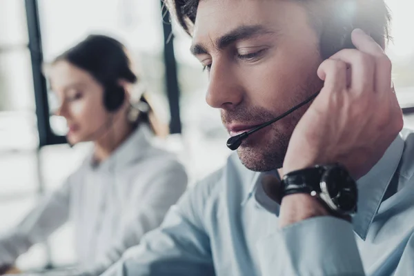 Guapo centro de llamadas gerente en auriculares con micrófono sentado en el lugar de trabajo, mientras que su colega sentado en segundo plano - foto de stock