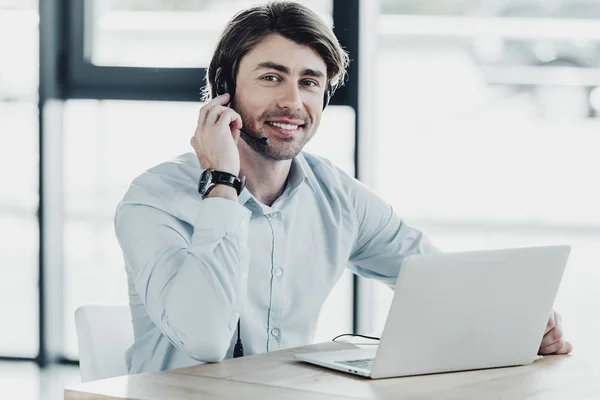 Trabalhador de call center sorridente com laptop olhando para a câmera enquanto sentado no local de trabalho — Fotografia de Stock