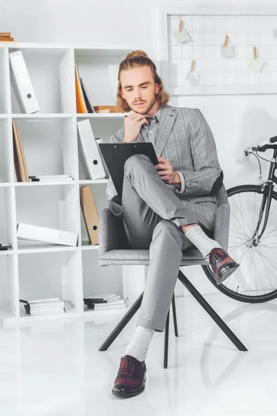 Nachdenklicher Geschäftsmann blickt auf Klemmbrett und sitzt im Stuhl — Stockfoto