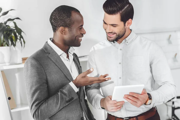 Sonrientes hombres de negocios afroamericanos y caucásicos hablando en la oficina y usando tableta - foto de stock
