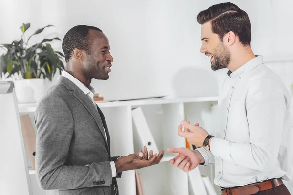 Sonrientes hombres de negocios afroamericanos y caucásicos hablando en la oficina - foto de stock