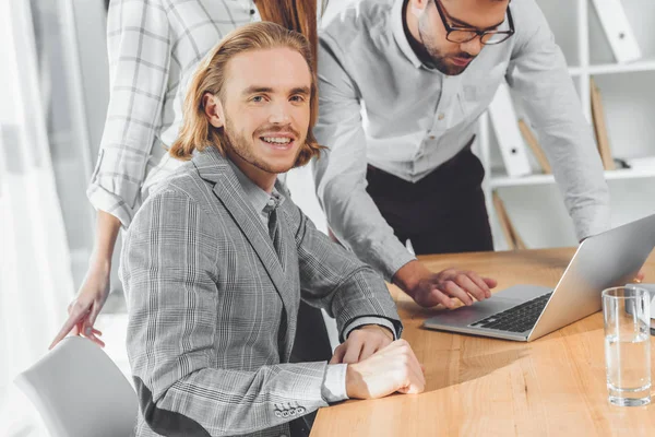 Hombre sonriendo a la cámara mientras que otras personas mirando el ordenador portátil en la mesa - foto de stock