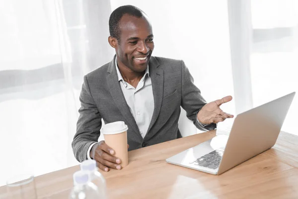 Hombre africano sonriente con taza de café sonriendo mientras mira en el portátil en la mesa - foto de stock