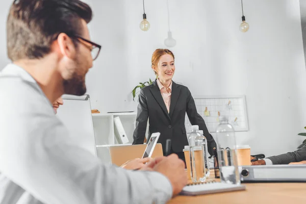Бізнес-леді в офіційному костюмі стоїть проти столу і розмовляє з командою в офісному просторі — стокове фото