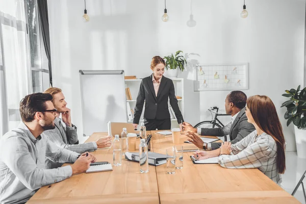 Businesswoman in formal suit standing against table and speaking to team at office space — Stock Photo