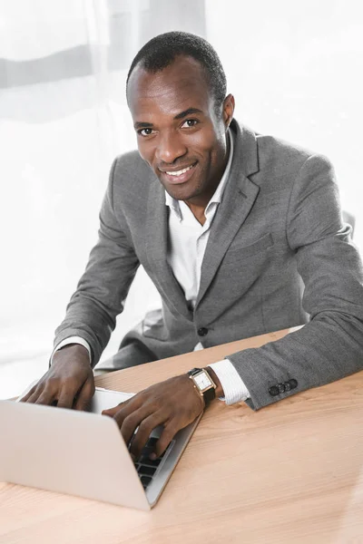 Hombre africano sonriente mirando a la cámara mientras usa el ordenador portátil en la mesa - foto de stock