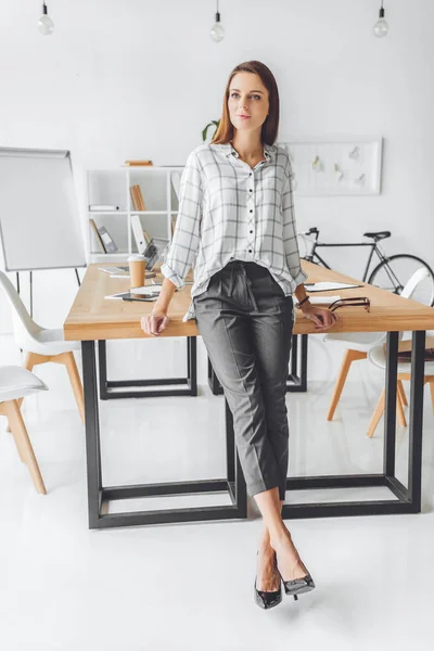Жінка в сорочці спирається на стіл і дивиться в офісний простір — стокове фото