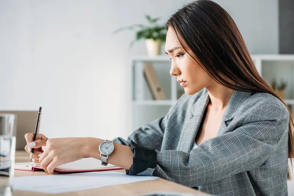 Vista laterale di grave attraente donna d'affari asiatica scrivere qualcosa per notebook in ufficio — Foto stock