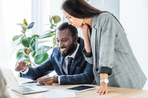 Lächelnder afrikanisch-amerikanischer Geschäftsmann zeigt asiatischer Geschäftsfrau auf etwas am Laptop — Stockfoto