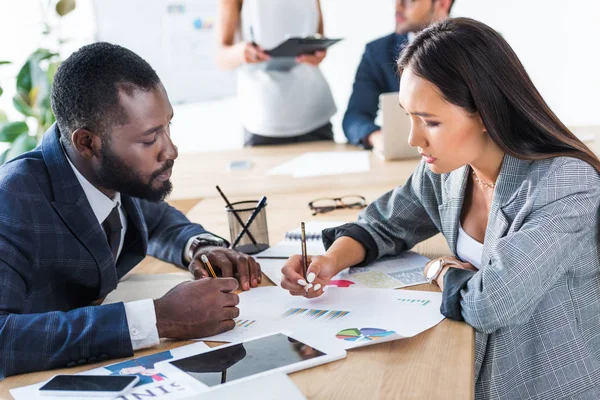 Side view of young multicultural businesspeople holding pencils and looking at documents in office — Stock Photo