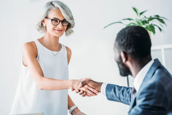 Hombre de negocios afroamericano y alegre mujer de negocios caucásica estrechando la mano en la oficina - foto de stock